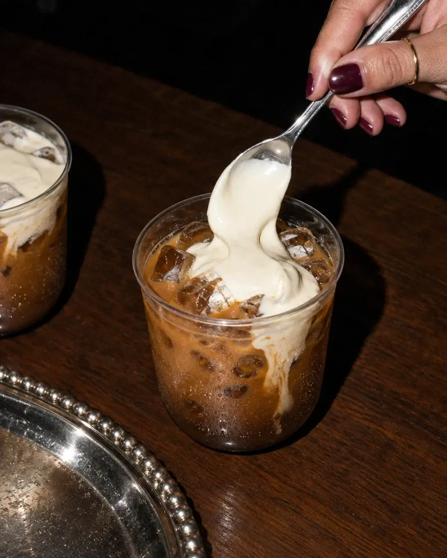 A spoonful of cream folded onto an iced phin coffee, beside a beaded silver tray.